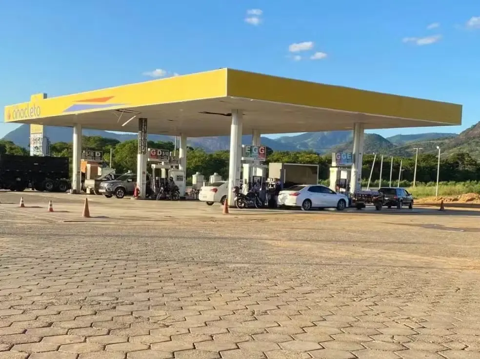 Imagem de um posto de combustíveis com bomba de gasolina em um dia ensolarado. Vários veículos estão estacionados no local, com montanhas ao fundo.