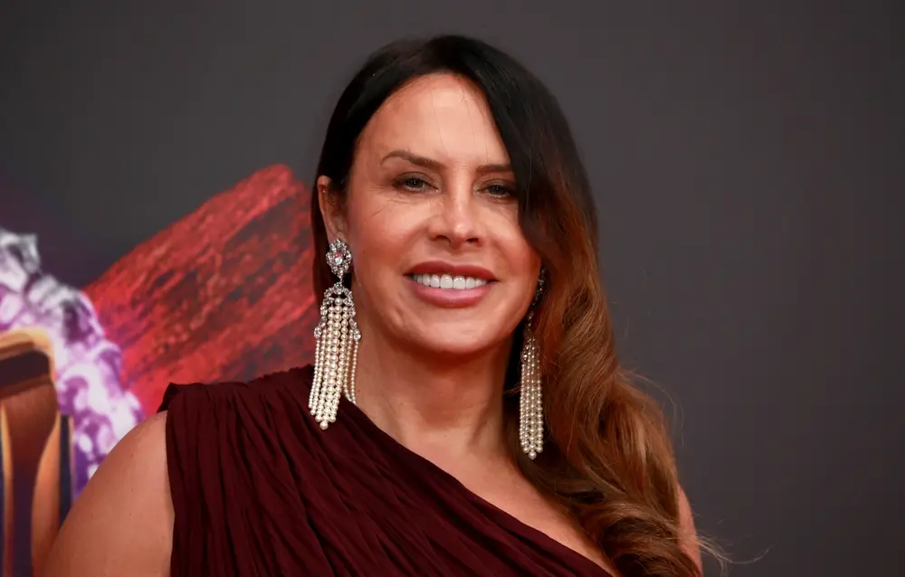 Karla Sofía Gascón é uma atriz espanhola trans de cabelos longos castanhos. Na foto, ela usa longos brincos de pérolas e vestido cor vinho