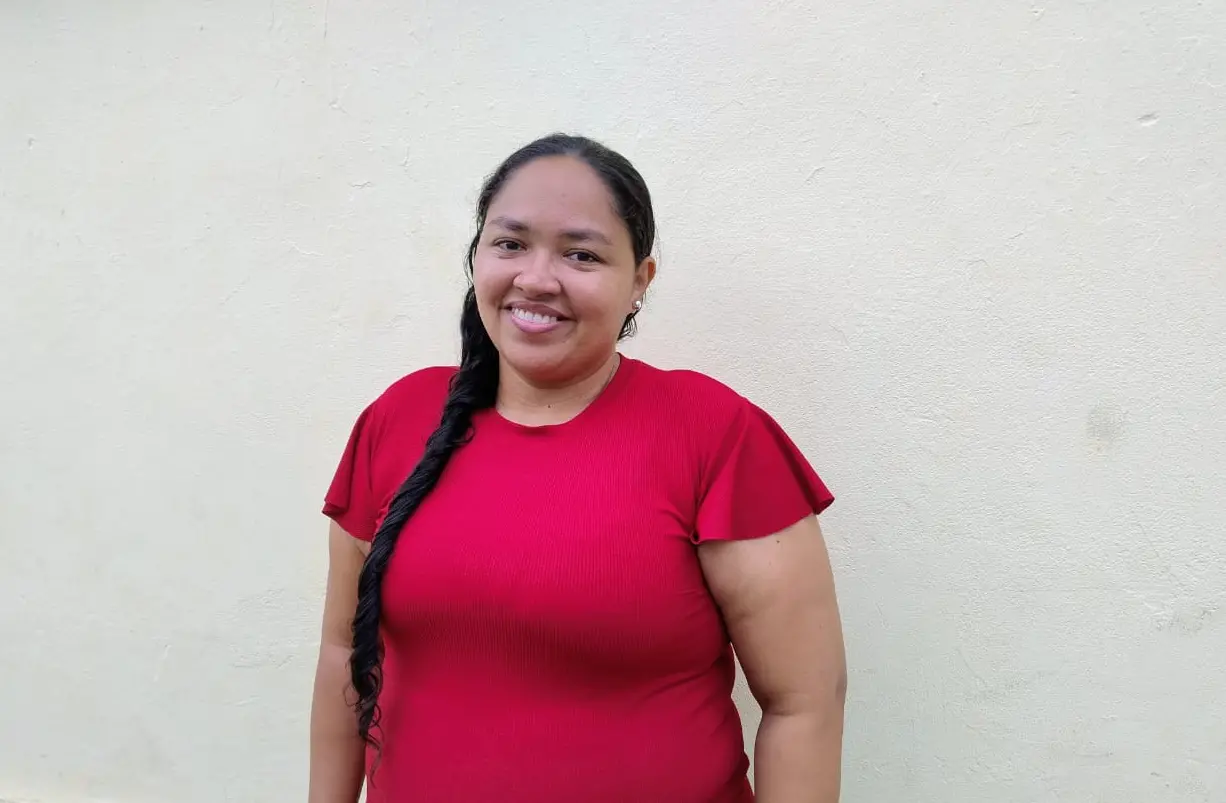 Jéssica sorrindo para a foto. Ao fundo, um muro amarelo. Ela veste uma camisa vermelha