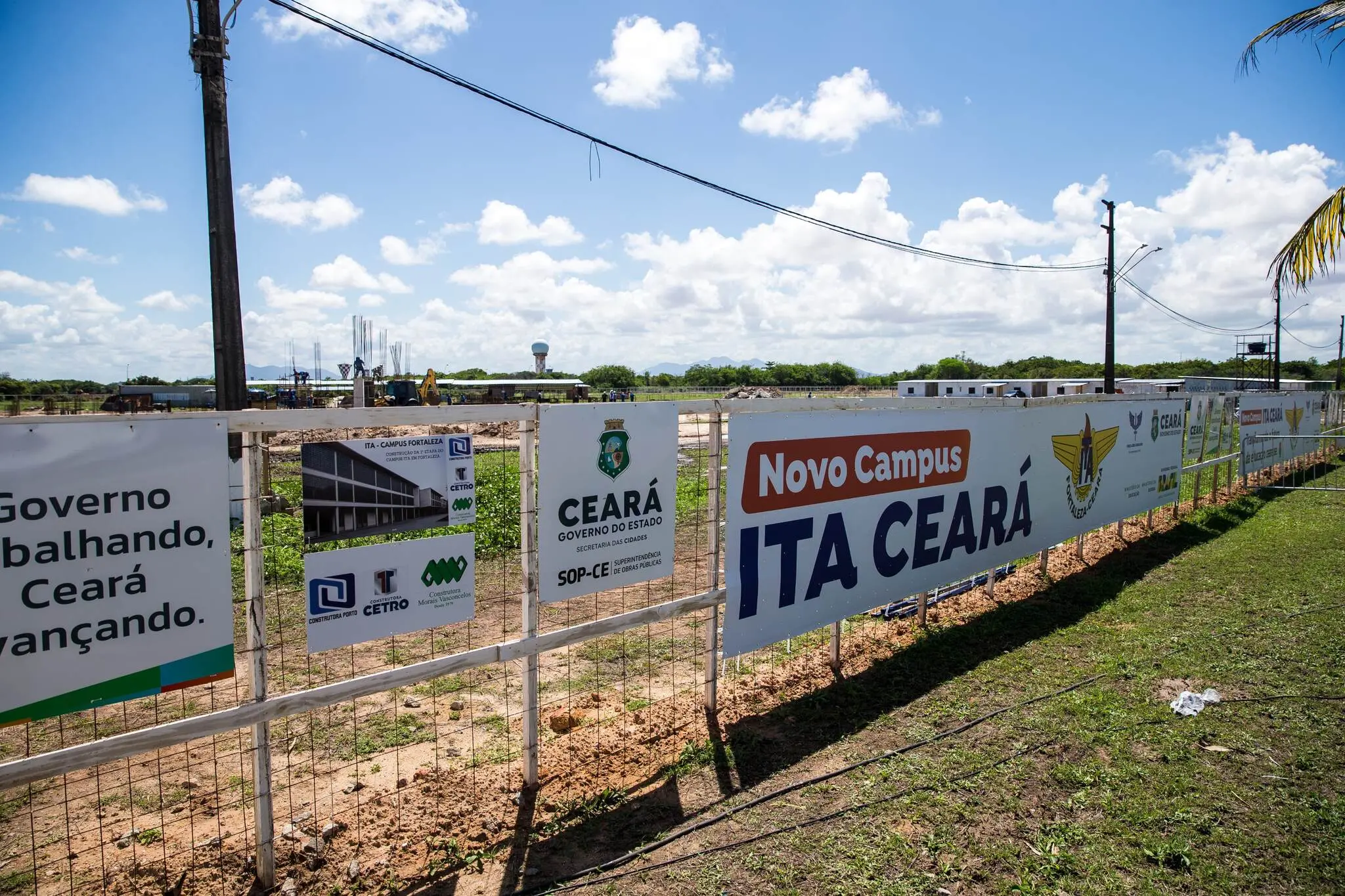 Vista ampla do canteiro de obras com cercas e placas indicando a construção do novo campus do ITA Ceará, céu azul e vegetação ao fundo.