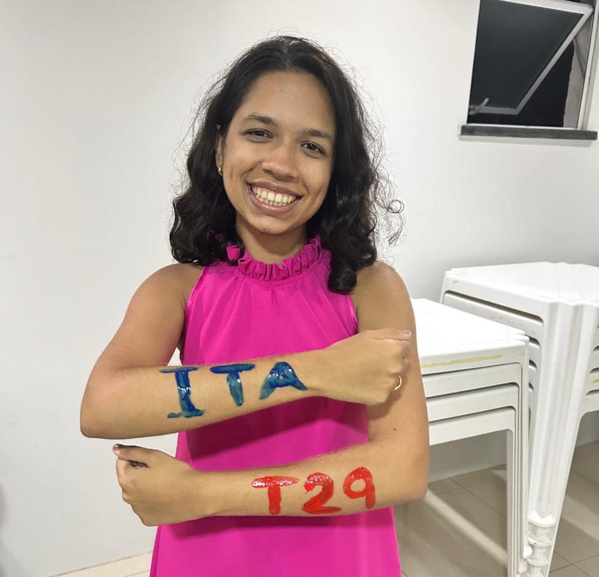 Marcela veste um vestido rosa e tem as palavras ITA e T29 escritas nos braços