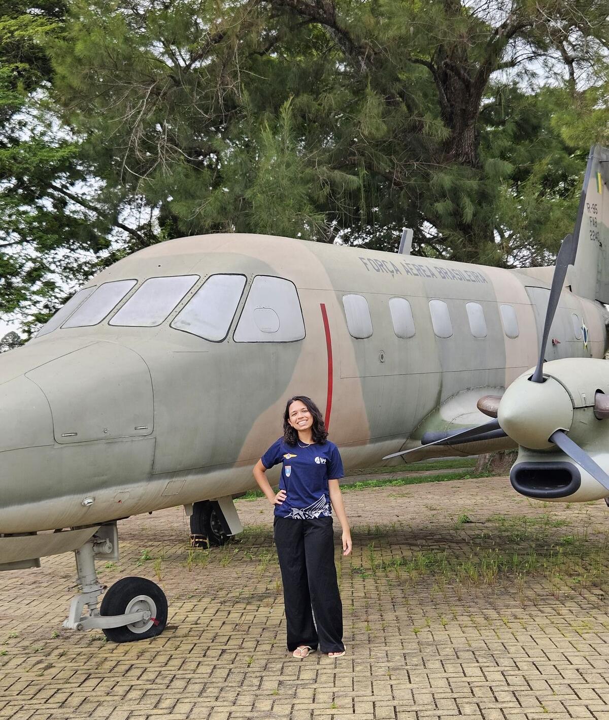 Marcela posa em frente a um avião verde militar da Força Aérea Brasileira