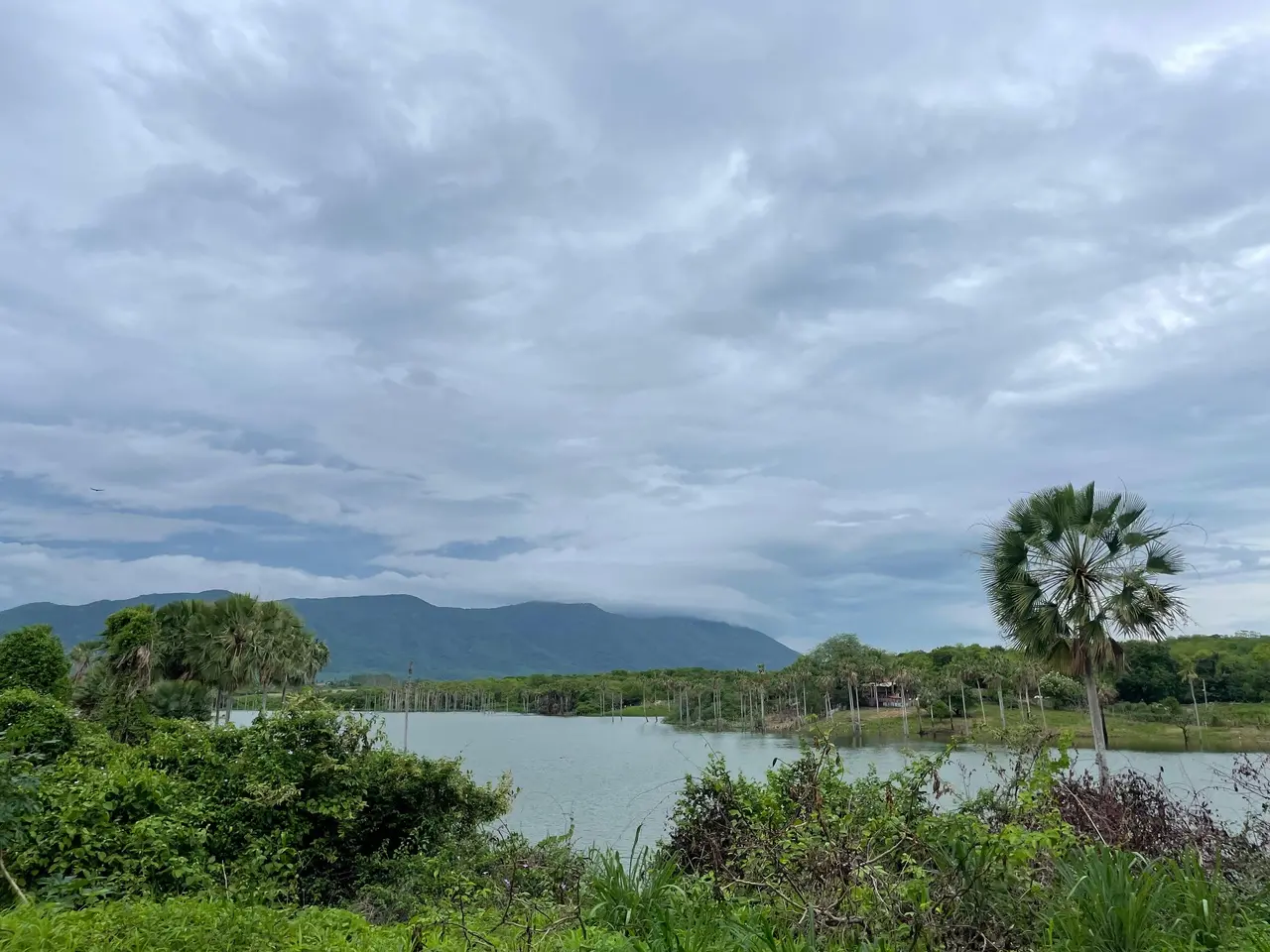 Paisagem com céu nublado, lago, vegetação, montanha ao fundo