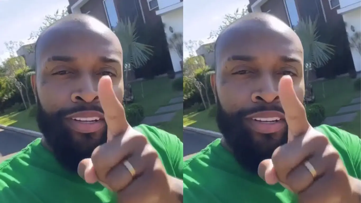 Foto de um homem com barba sorrindo, usando uma camiseta verde e fazendo um gesto de alerta ao segurar o dedo indicador em direção à câmera, em um ambiente externo residencial com plantas ao fundo.
