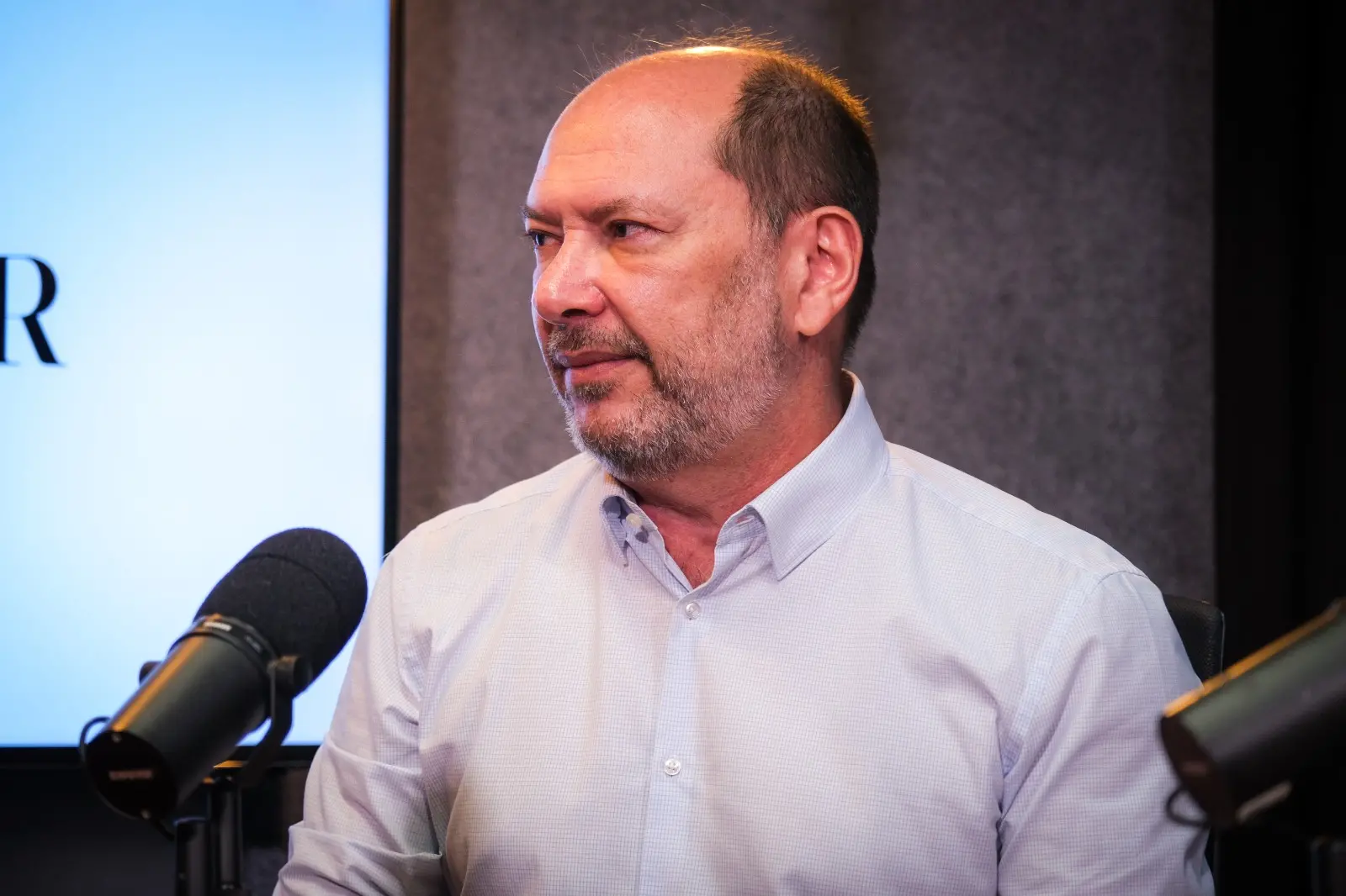 Homem branco calvo, com cabelos ralos castanhos nas laterais e pouca barba e bigode brancos, usando camisa social branca.
