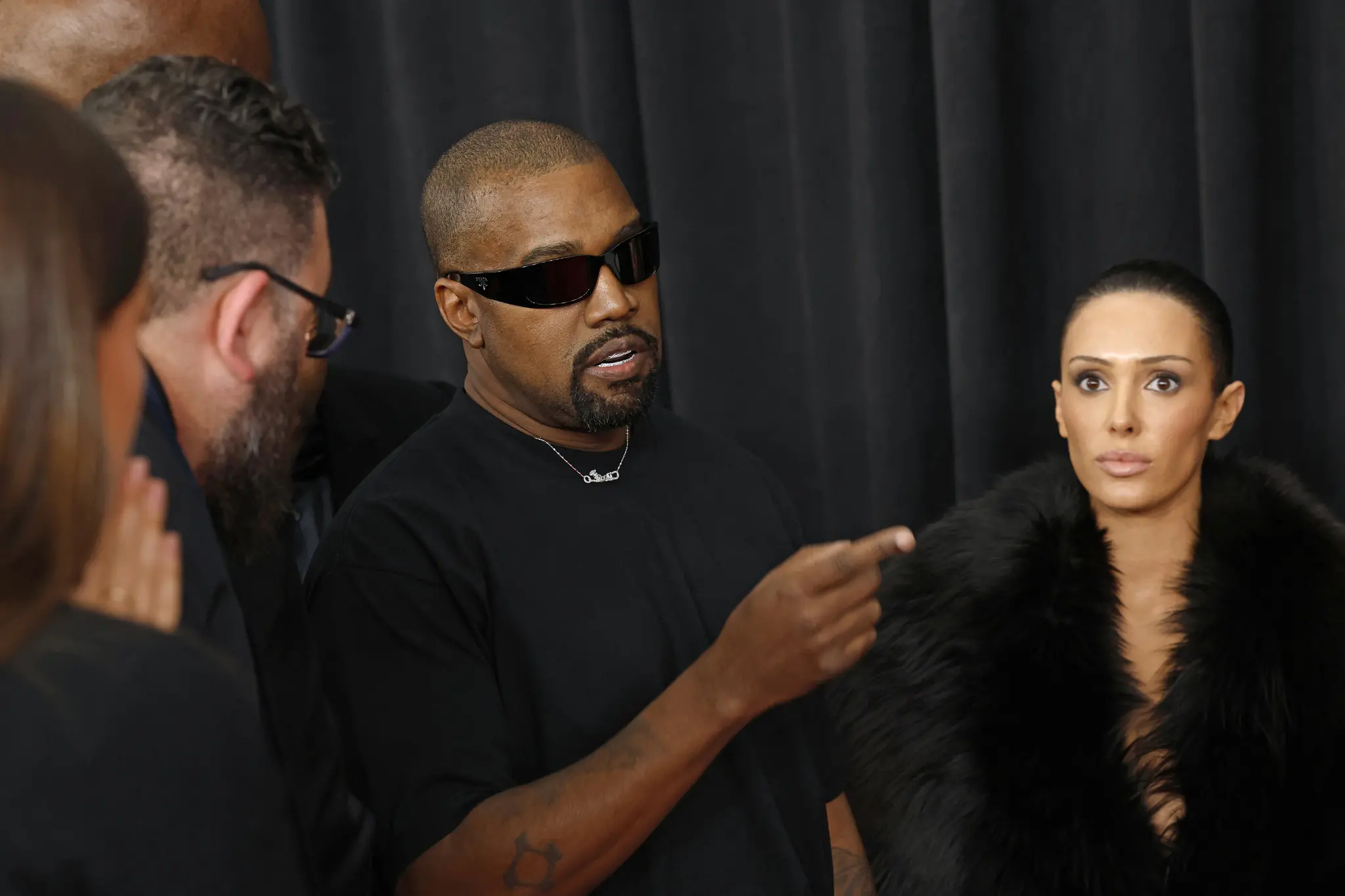 Kanye West falando, apontando com o dedo para frente, e a esposa, Bianca Censori, ao lado
