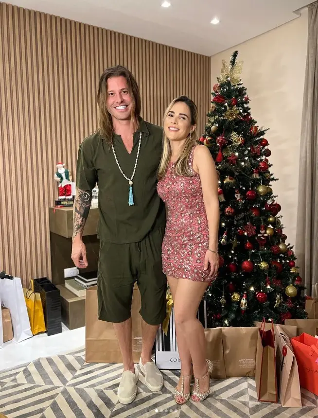 Foto de casal sorridente em pé ao lado de uma árvore de Natal decorada, com presentes e um ambiente festivo ao fundo.
