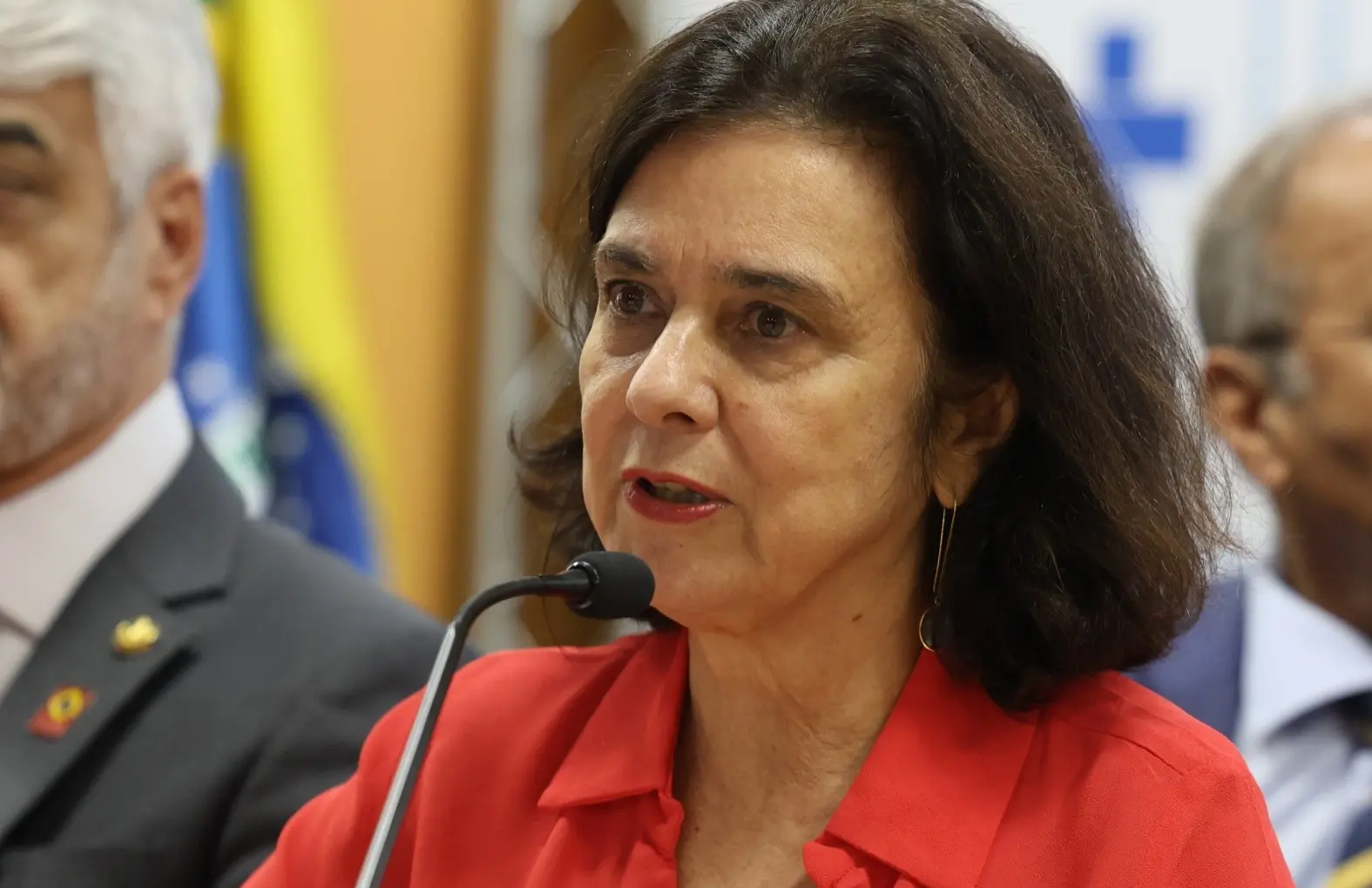 Nísia é uma mulher branca de cabelo tamanho médio liso e castanho escuro. Na foto, ela está falando ao microfone e usa camisa social vermelha