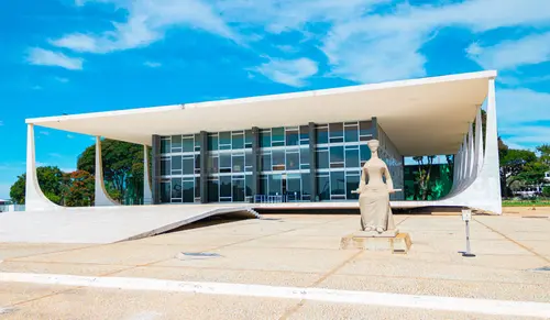 Imagem do Supremo Tribunal Federal com sua arquitetura moderna e a escultura simbólica em primeiro plano, em um dia ensolarado e céu azul.