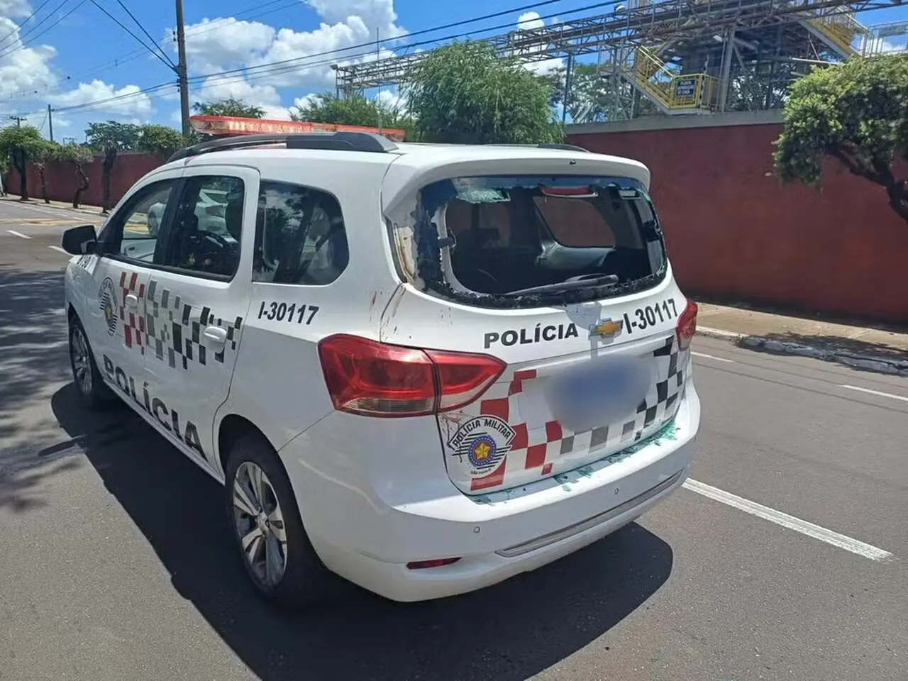 Imagem de uma viatura da polícia com janelas quebradas, estacionada em uma rua. O veículo apresenta marcas de danos e a placa de identificação é visível.