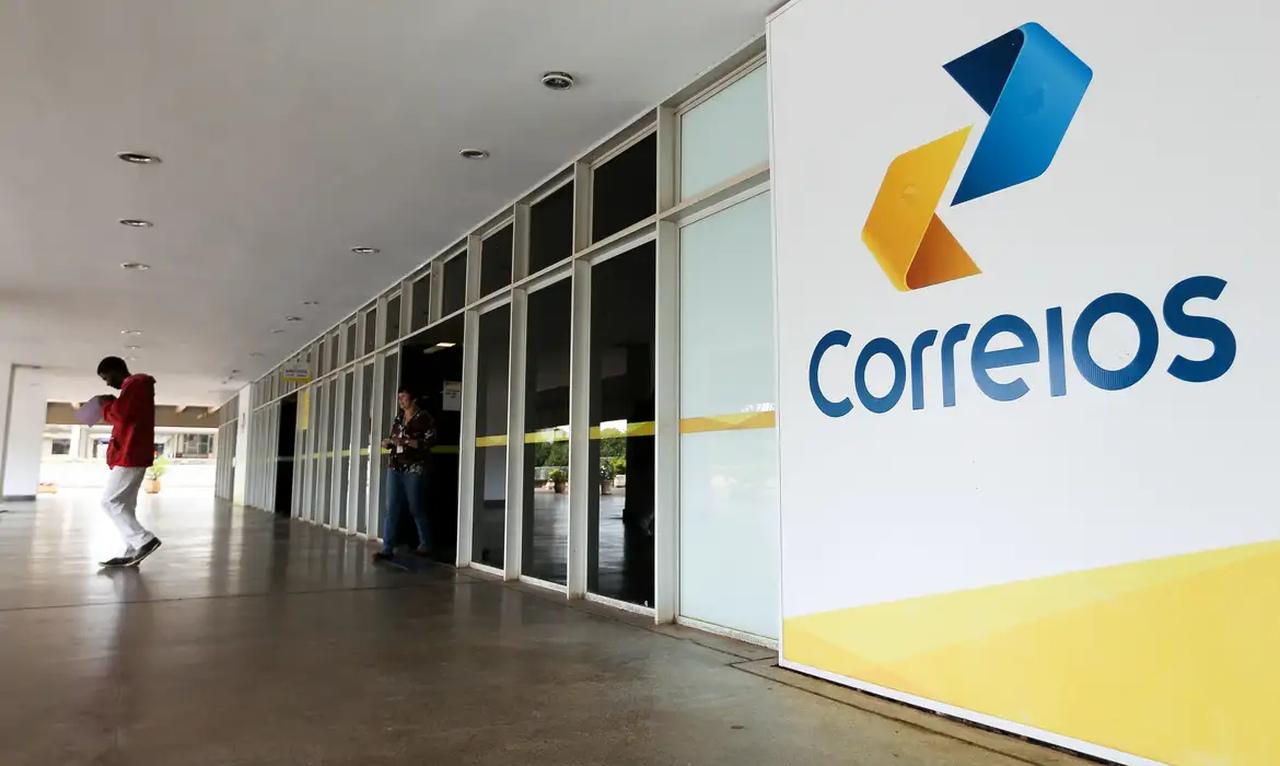 Imagem da entrada da agência dos Correios, com o logotipo visível, mostrando um ambiente comercial em um dia comum.