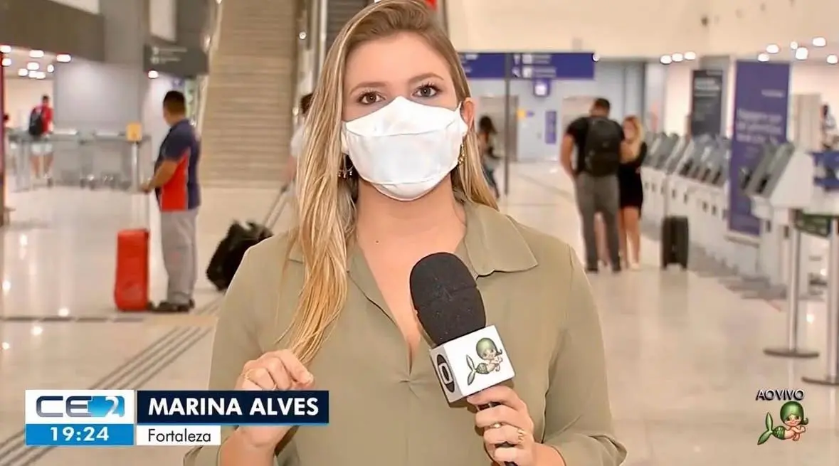 Marina Alves como repórter da TV Verdes Mares. Na imagem, ela está de camisa de gola polo marrom claro e usa máscara branca cobrindo o rosto