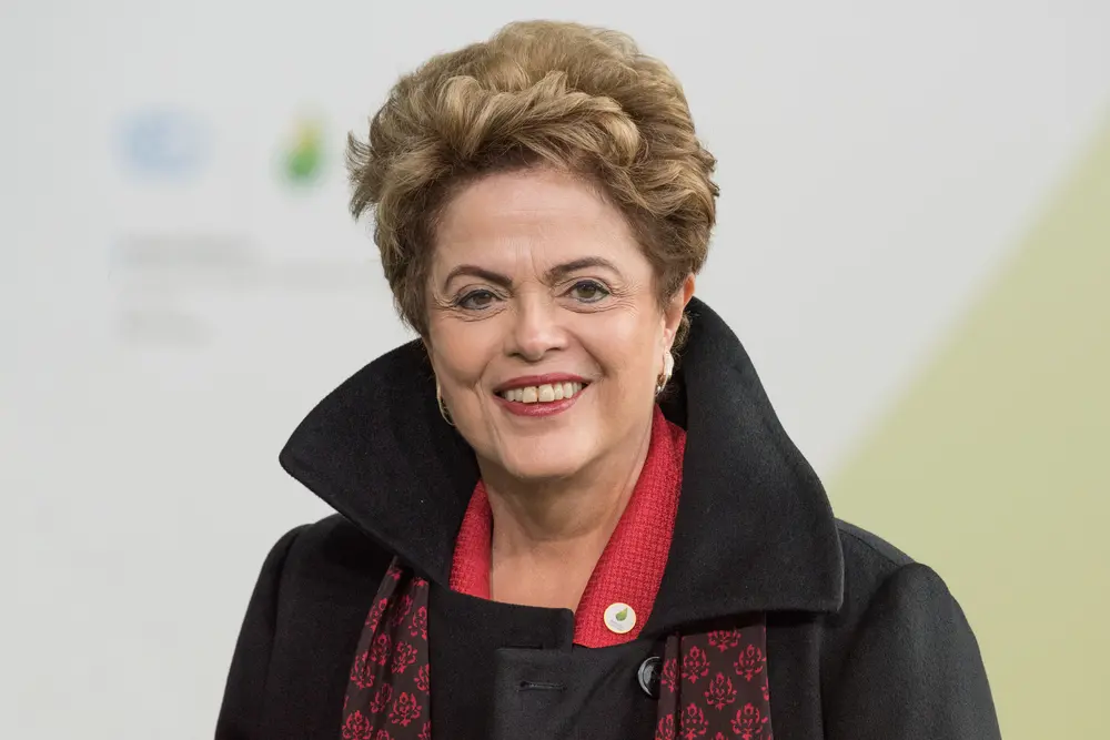 Dilma é uma mulher idosa de cabelo curto castanho. Na foto, ela está de casaco preto, sorrindo. Ela usa batom vermelho