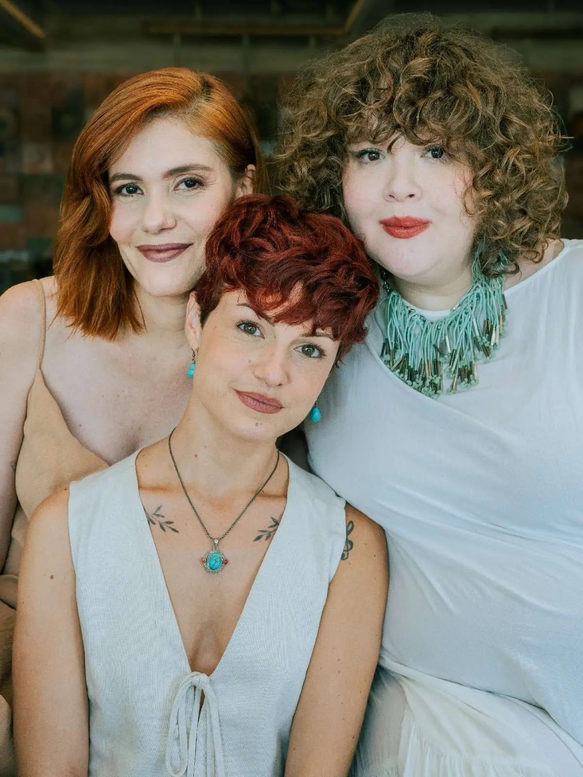 Foto de três mulheres sorrindo, em primeiro plano, com diferentes estilos de cabelo e roupas leves, em um ambiente iluminado.