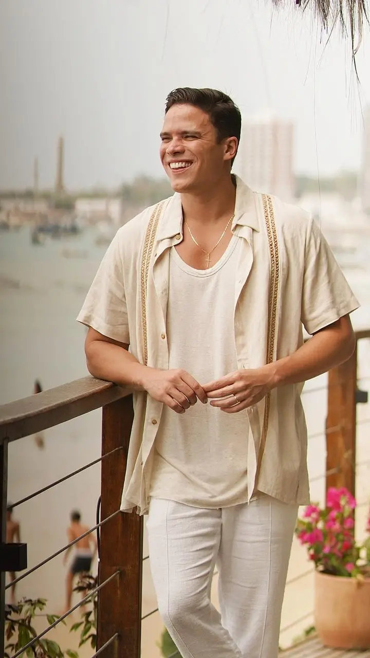 Foto de Marcos Lessa sorridente em um ambiente tropical, usando uma camisa clara e calças brancas, posando em uma varanda com vista para o mar.