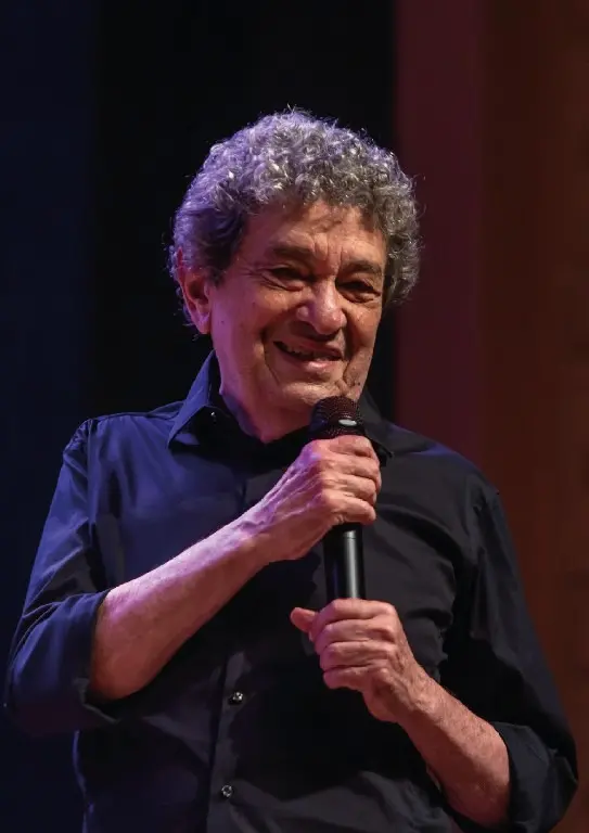 Foto de Fausto Nilo sorridente segurando um microfone, durante uma apresentação no palco. Ele possui cabelos grisalhos e usa uma camisa preta.