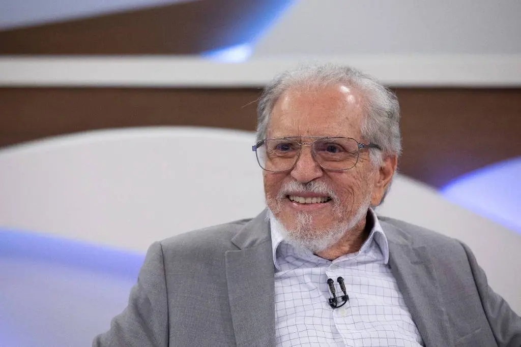 Imagem do apresentador Carlos Roberto de Nóbrega sorridente em um programa de TV, usando óculos e um terno cinza. Ele transmite simpatia e carisma, ideal para conteúdos sobre entrevistas e personalidades da televisão.
