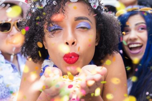 Foto de uma jovem mulher preta, com cabelo crespo e maquiagem colorida, exibindo um sorriso enquanto sopra confete e serpentina em uma festa animada, rodeada de amigos.