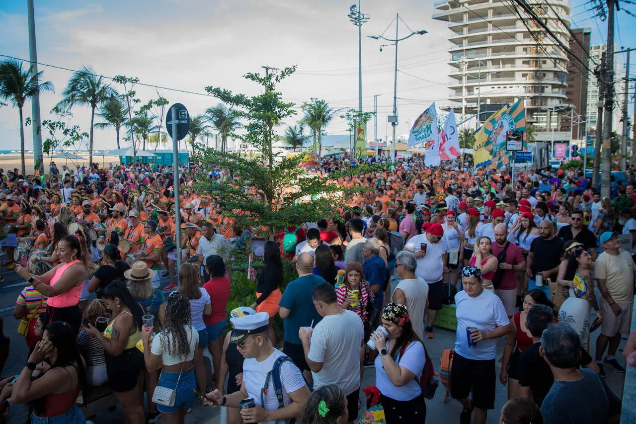 Multidão animada durante o carnaval na praia de Fortaleza, com pessoas fantasiadas e bandeiras, em um ambiente festivo e alegre.