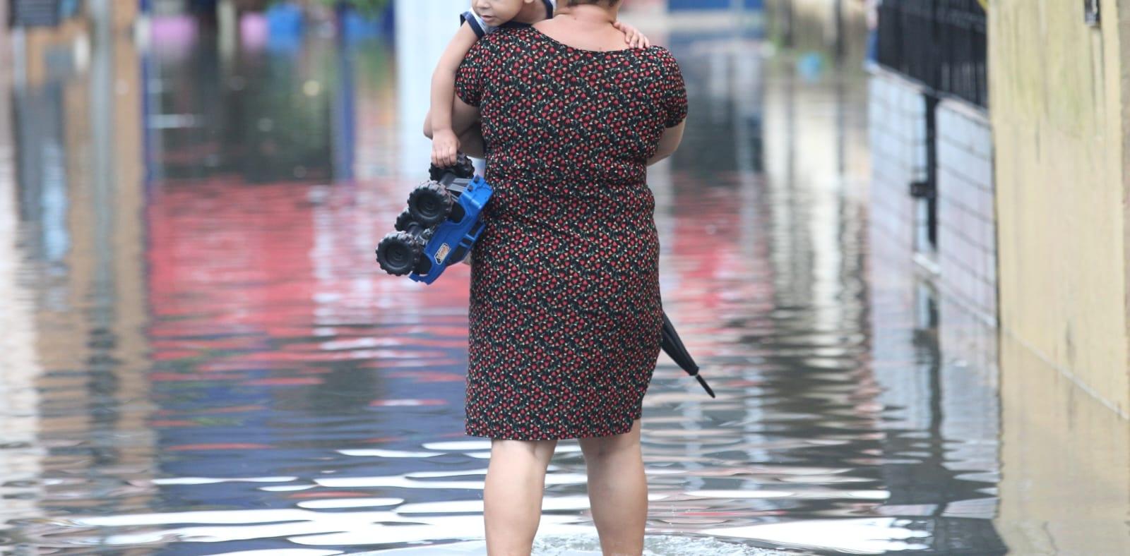 Mulher caminha com criança no braço em rua alagada em Fortaleza