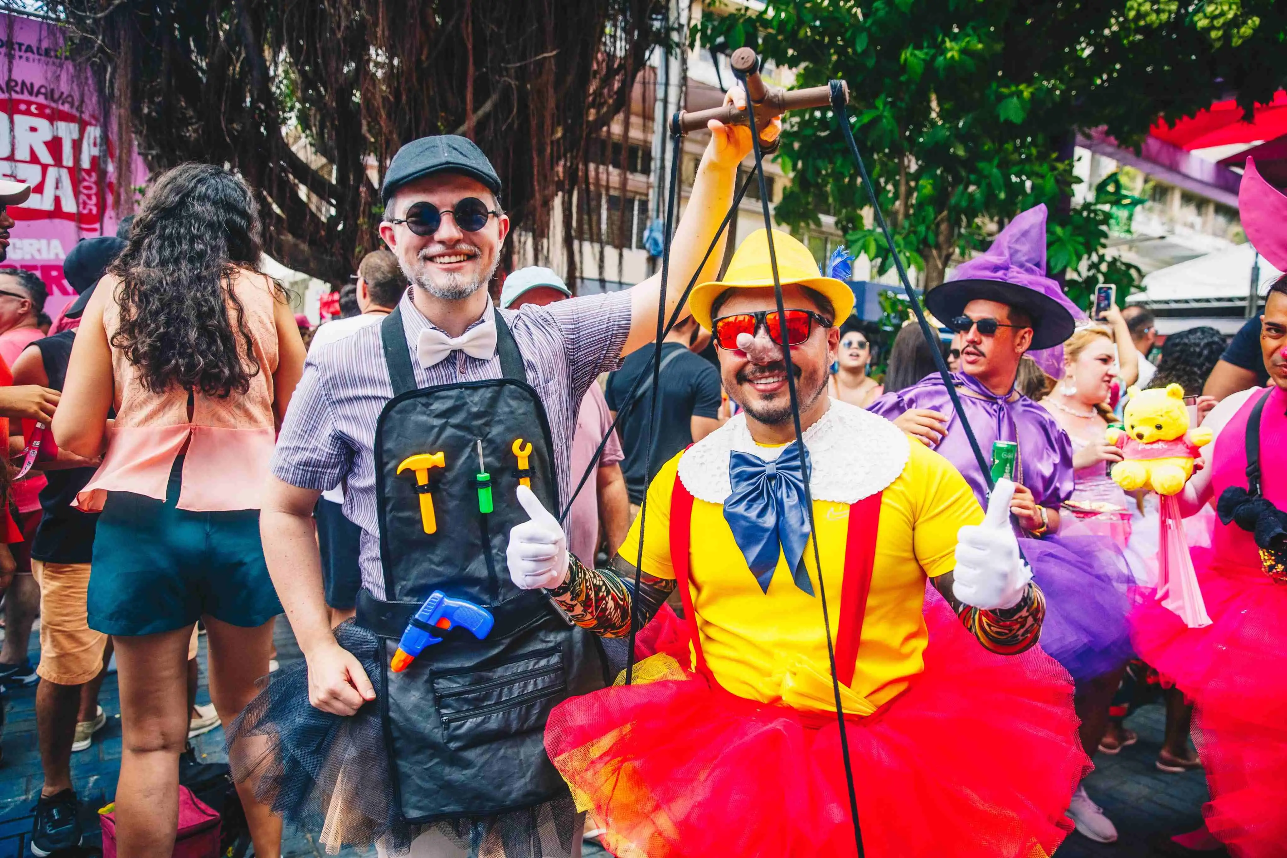 Foto de pessoas fantasiadas em uma festa de carnaval, um homem vestido de palhaço colorido e um amigo em trajes divertidos, celebrando ao ar livre em ambiente festivo.