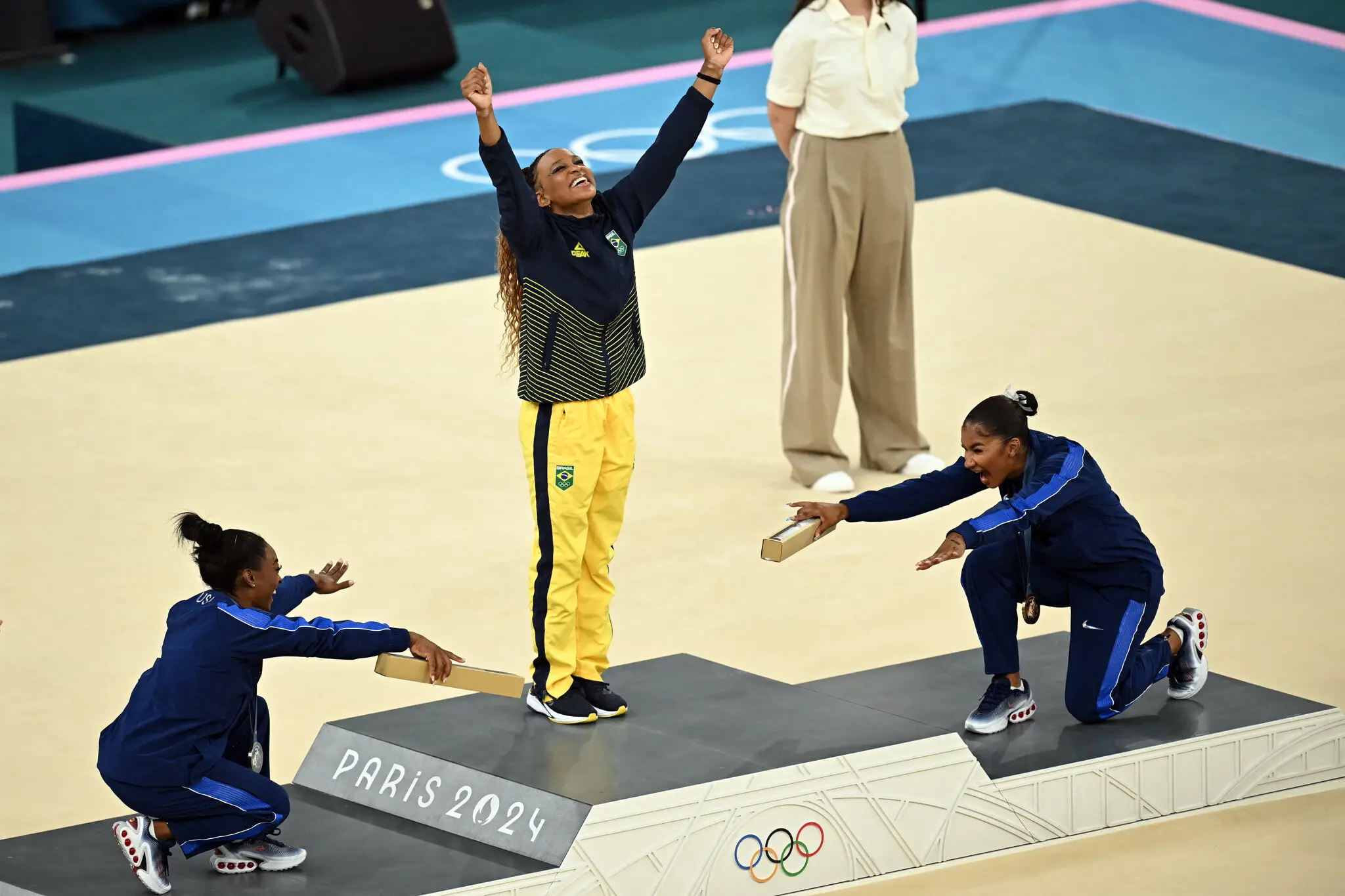 Foto da Brasileira Rebeca Andrade sendo reverenciada pelas norte-americanas Simone Biles e Jordan Chiles após levar o ouro em Paris 2024