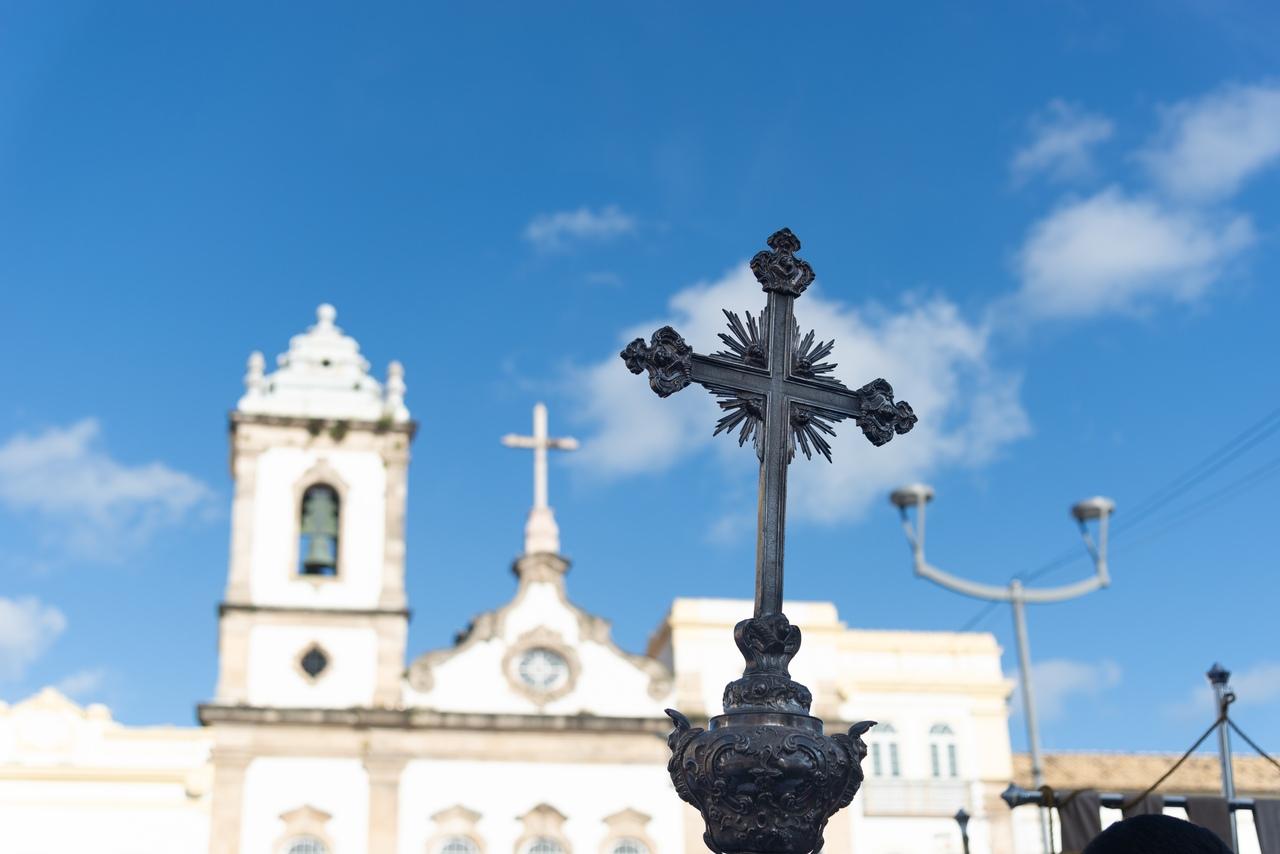 Cruz de metal em destaque com céu azul ao fundo e igreja ao fundo, representando um ponto turístico religioso.