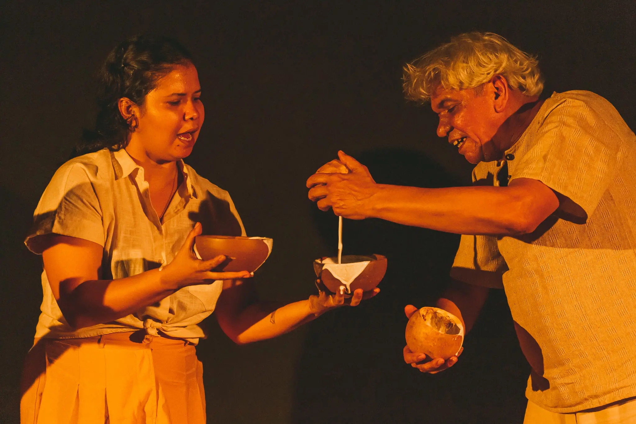 Cena de uma apresentação teatral que retrata a interação entre dois personagens, um homem e uma mulher, utilizando recipientes e ingredientes na cena.