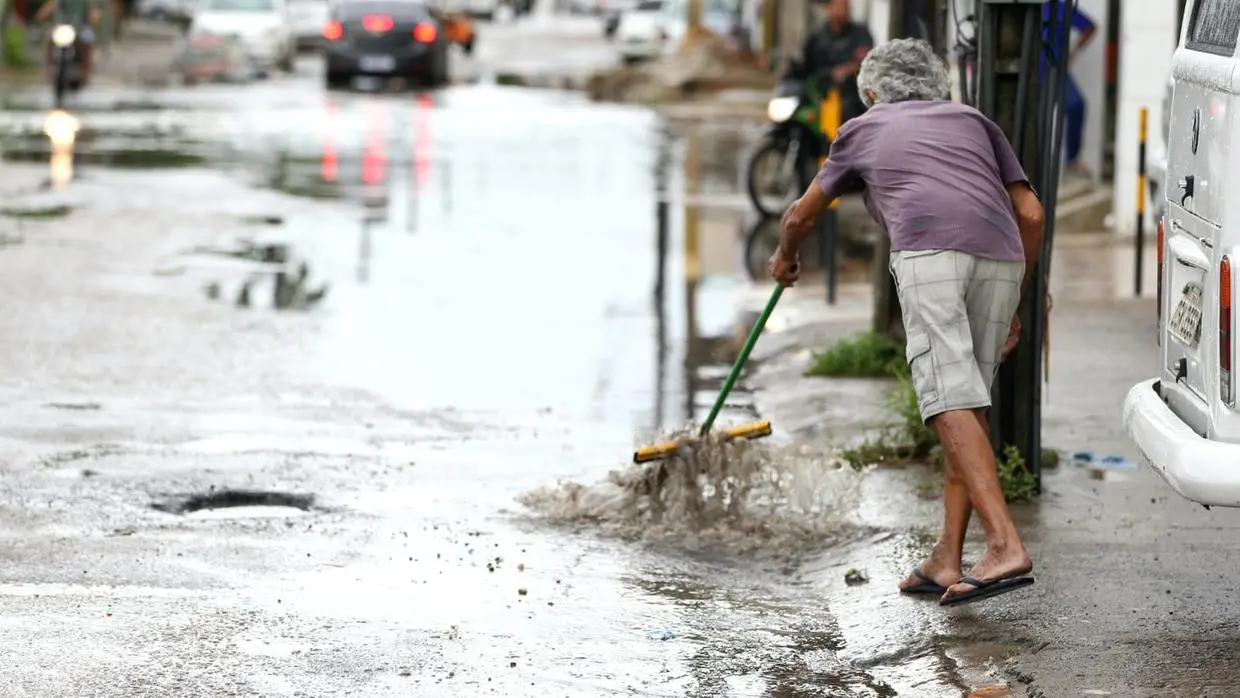 Imagem de homem idoso usando rodo para tenta escoar água das chuvas em frente de casa em Caucaia