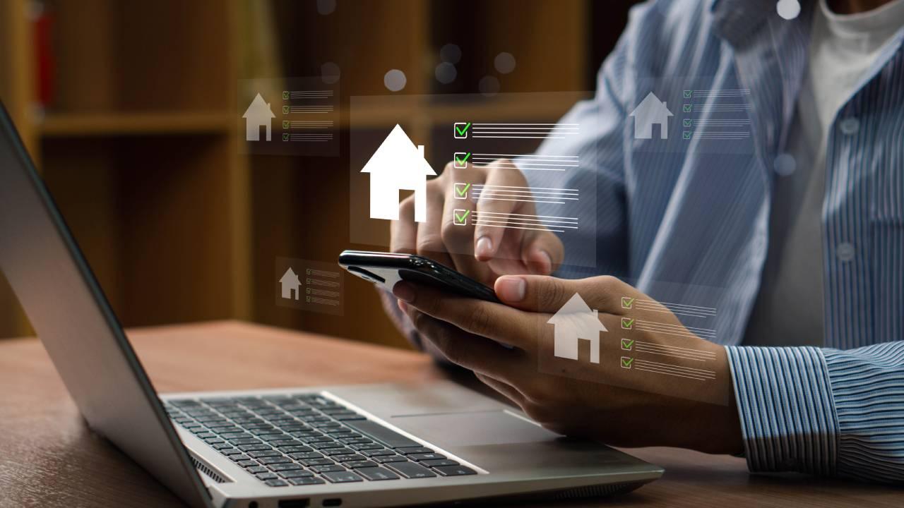 Homem segura celular em frente a computador e arte mostra o desenho de uma casa