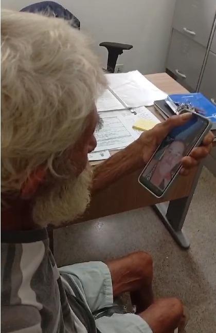 Idoso usando smartphone para videochamada enquanto está sentado em uma mesa de escritório, foco na interação digital e no cotidiano.