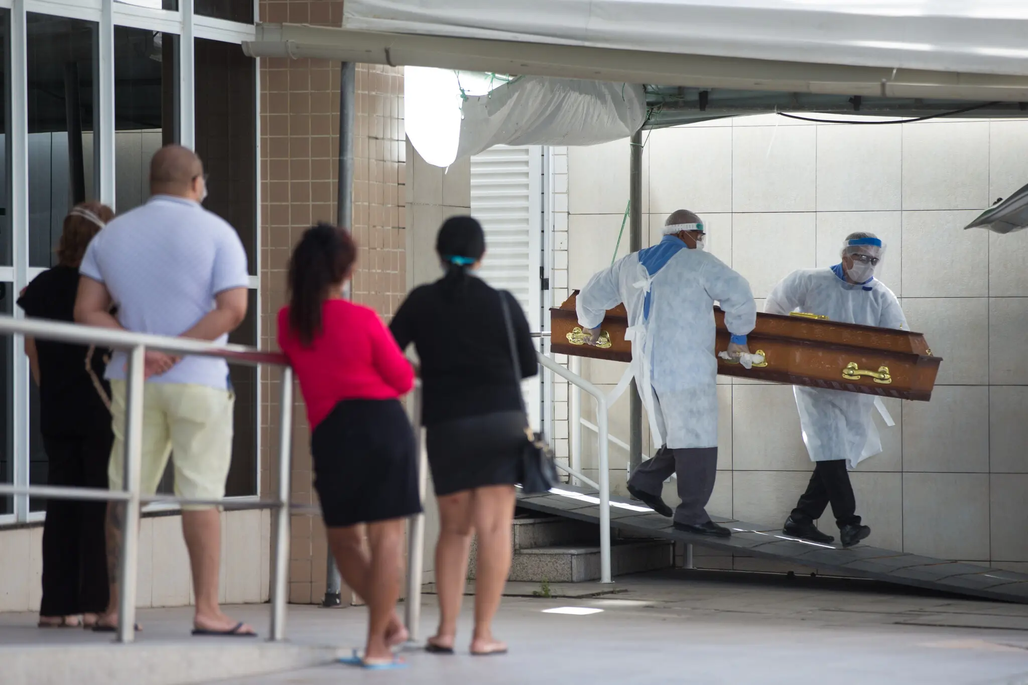 Pessoas na frente de hospital de Fortaleza veem profissionais da saúde com roupa especial para evitar contágio colocando caixão em carro funerário