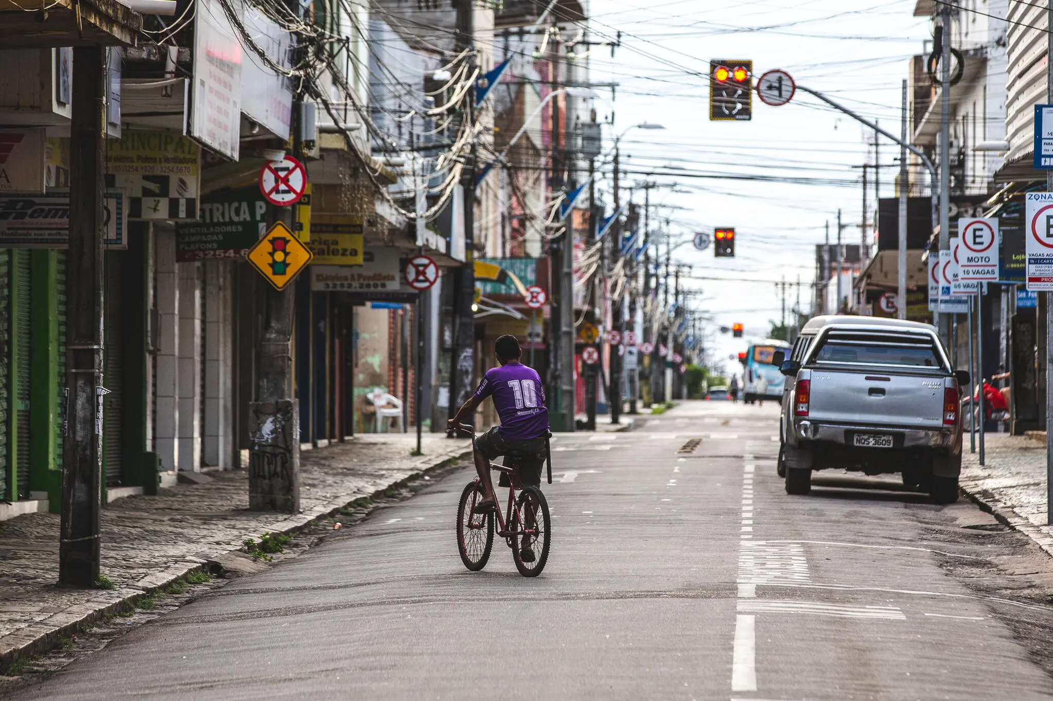 Rua do centro com pouco movimento, apenas um ciclista de costas e poucos veículos estacionados