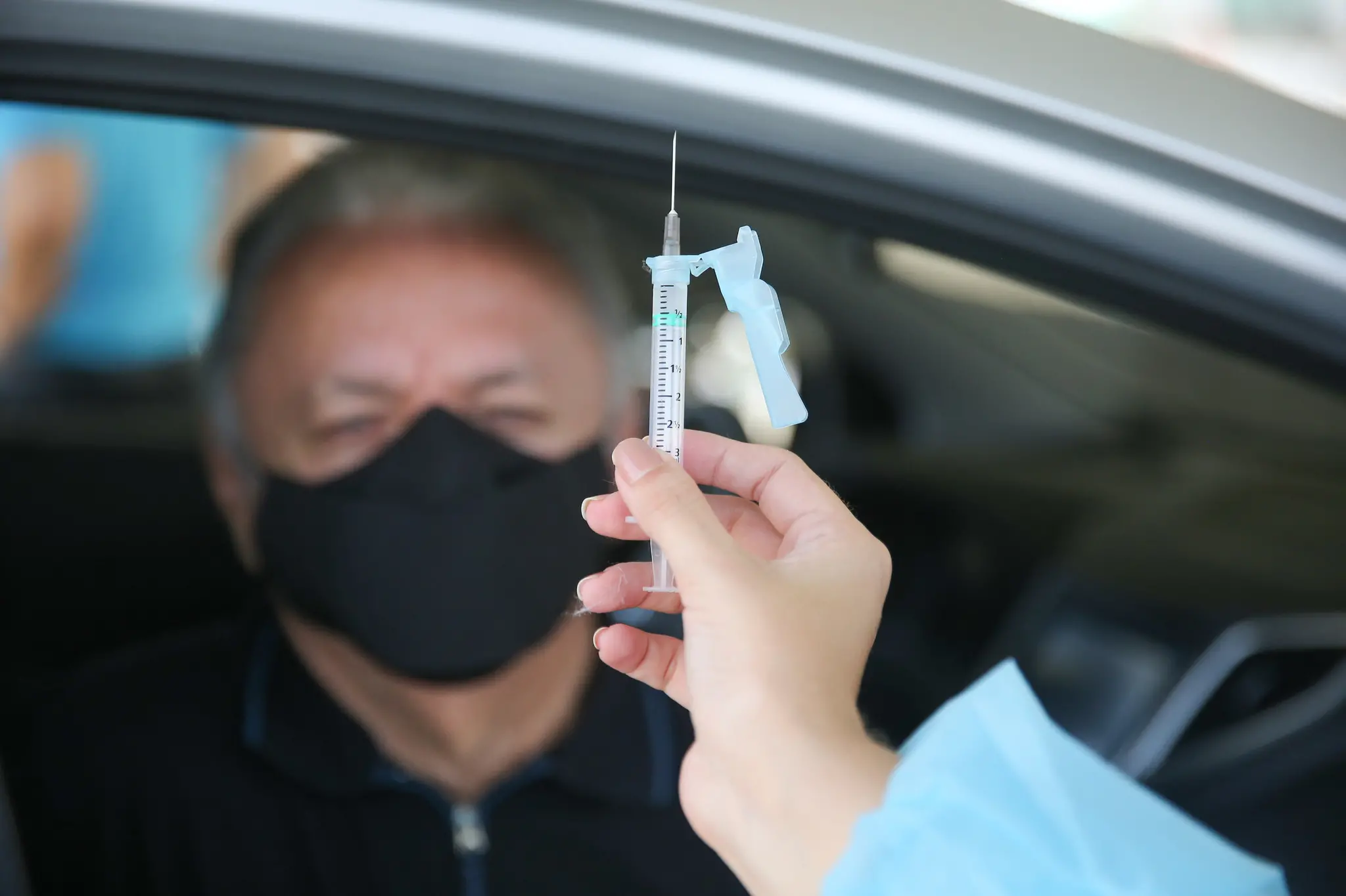 Mão de profissional da saúde segurando uma seringa com vacina. Ao fundo, rosto desfocado de um idoso usando máscara