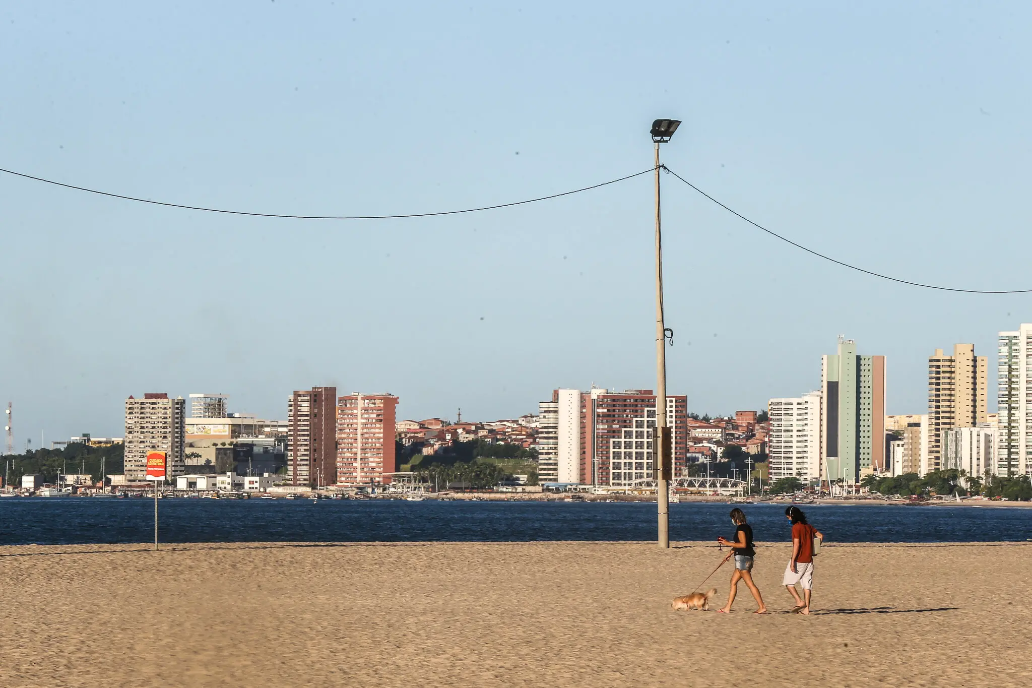 Vista de uma praia com apenas um casal passeando usando máscara e com cachorro