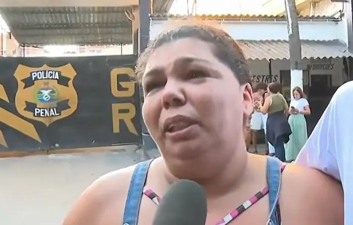 Débora é uma mulher de meia idade, branca. Na foto, ela está de cabelo amarrado em um rabo de cavalo e com o semblante triste enquanto concede uma entrevista em frente ao presídio no Rio de Janeiro