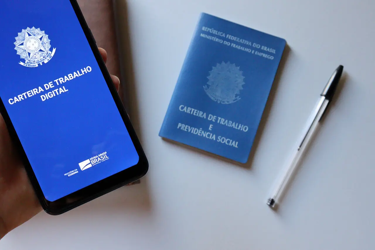 Imagem da Carteira de Trabalho Digital sendo exibida em um smartphone, ao lado da Carteira de Trabalho física e uma caneta, representando a modernidade no acesso a documentos trabalhistas.