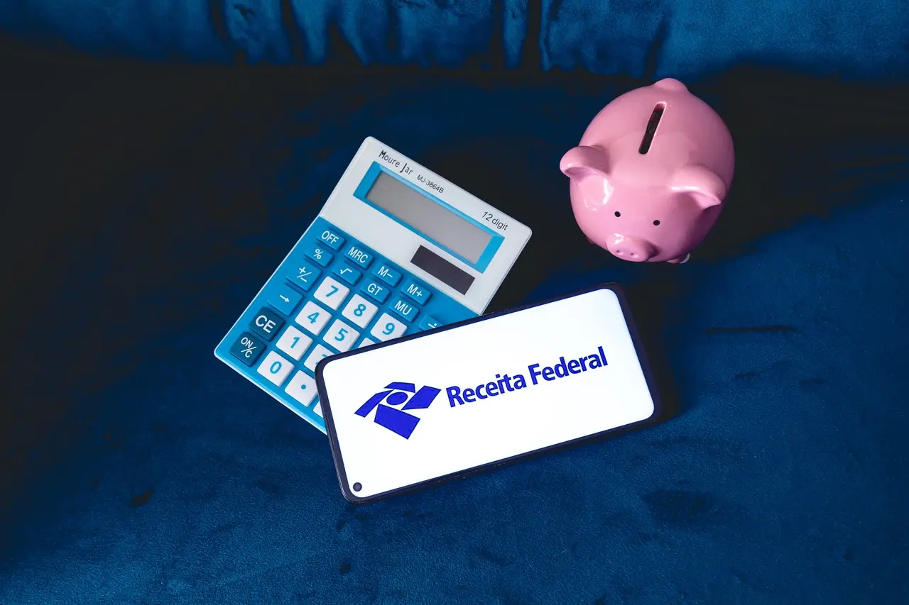 Imagem com uma calculadora, um cofre de porquinho rosa e a logo da Receita Federal, simbolizando finanças e impostos.