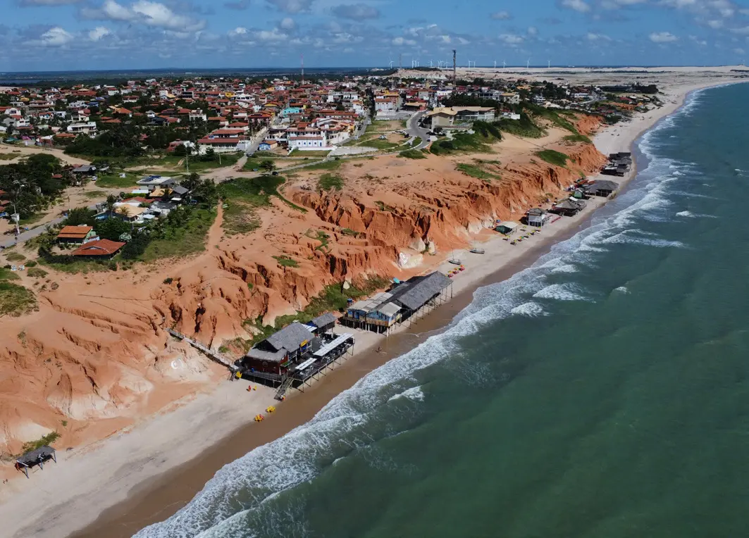 Erosão costeira na praia de Canoa Quebrada, Aracati, em 2024, mostrando uma estreita faixa de areia onde estão posicionadas as barracas de praia e, ao fundo, a vila de Canoa Quebrada