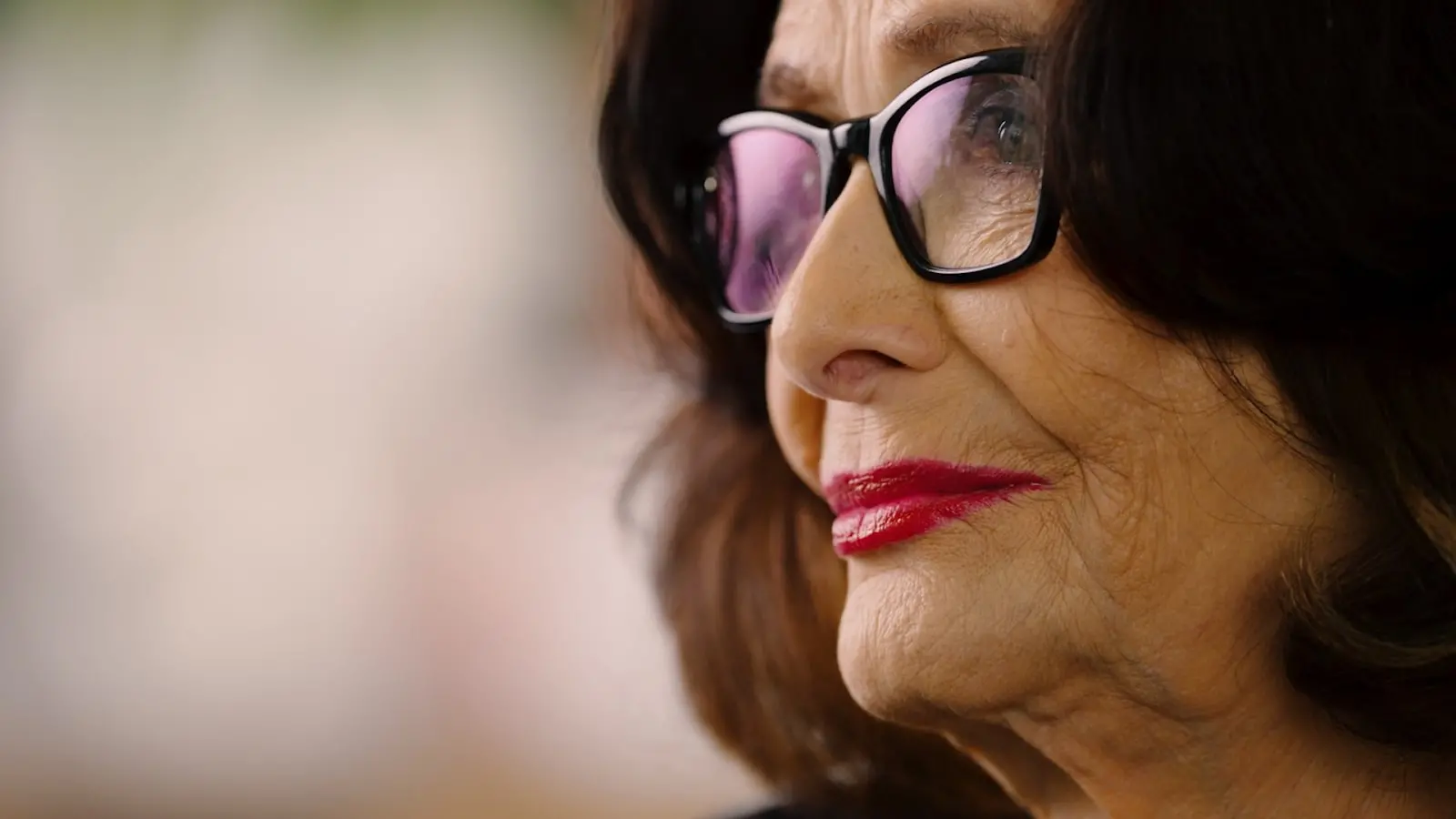 Retrato de uma mulher mais velha com cabelo escuro e óculos. Ela possui lábios vermelhos e uma expressão serena. A imagem destaca a beleza e a sabedoria da idade.