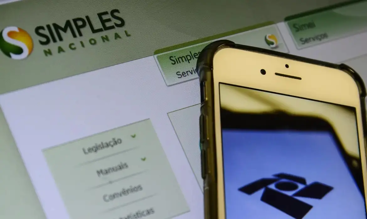 Imagem mostra tela de computador com página do Simple Nacional, serviço que emite nota fiscal, aberta sob celular com logo na receita federal na tela. MEIs terão nova regra para emissão de nota fiscal em abril; vejo o que muda