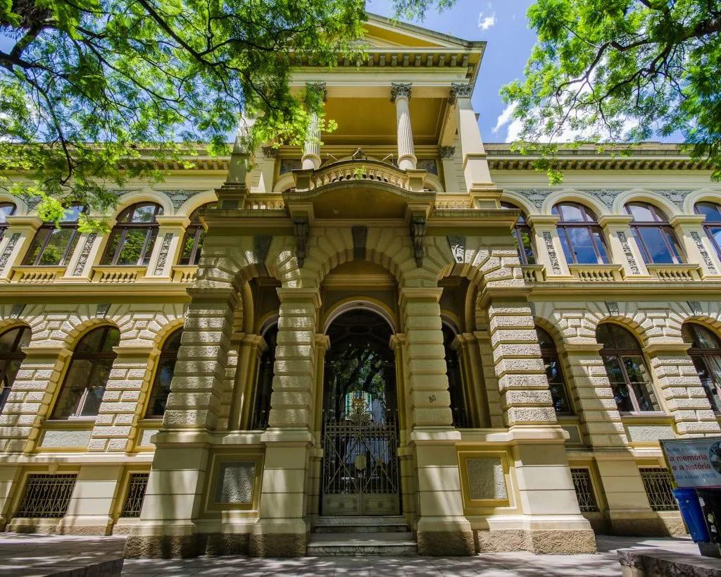 Antiga construção com arquitetura clássica, mostrando detalhes de colunas e arcadas, rodeada por árvores verdes, ideal para amantes da arquitetura histórica.