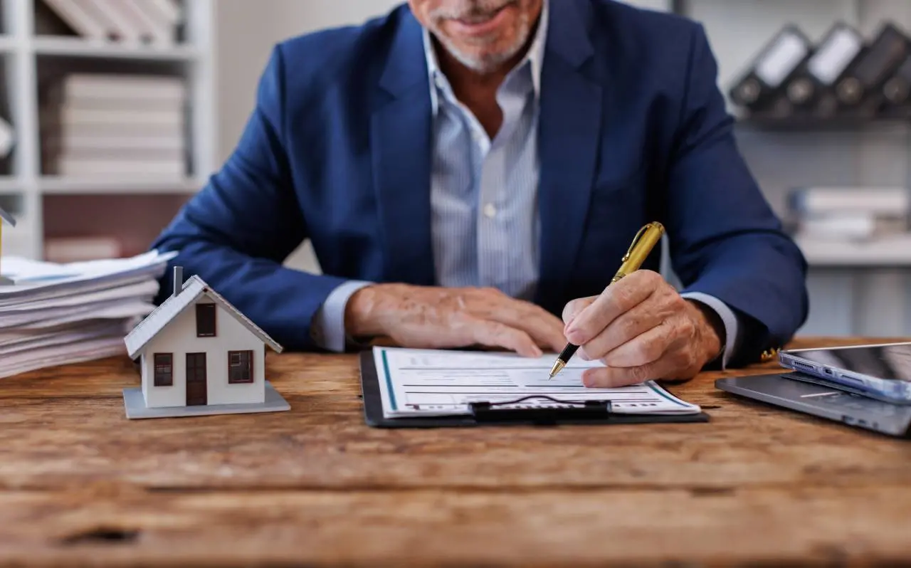 Home de blazer assina documento em mesa com maquete de casa ao lado