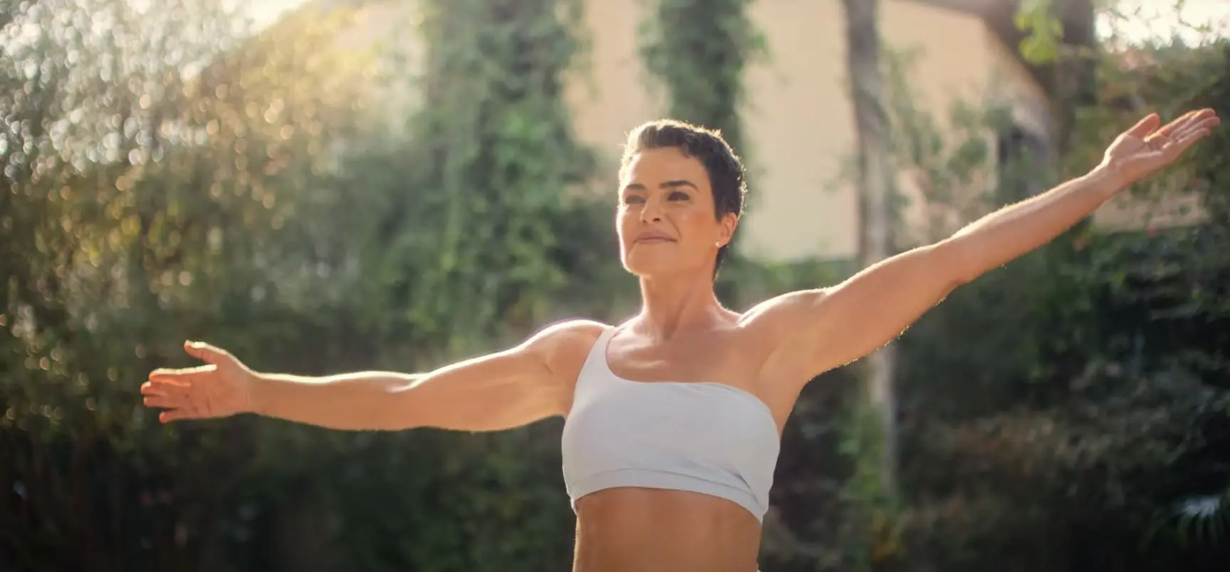 Ana Paula Arósio sorrindo vestindo roupa branca, de braços abertos, e em frente a cenário de céu azul e árvores