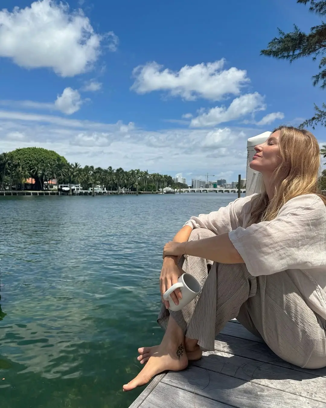 Gisele bundchen sentada, de olhos fechados, ao lado de uma lagoa. ela veste trajes claros e está de cabelos soltos