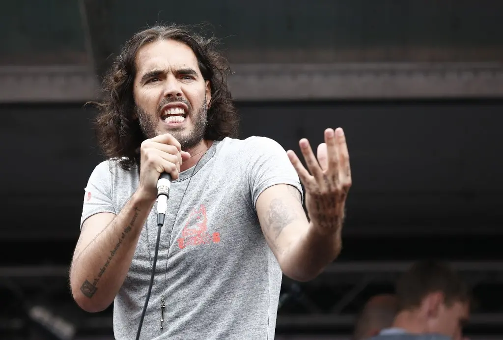 Russell Brand se apresenta emocionado em um palco, falando ao microfone e gesticulando, transmitindo sua mensagem para a plateia.