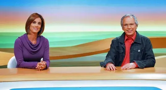 Helen Martins e Nelson Araújo apresentam o Globo Rural, com a bacada a frente de ambos e um fundo de estúdio atrás, com cores vibrantes