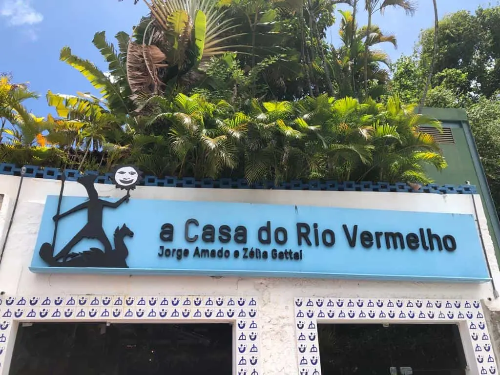 Imagem da fachada da Casa do Rio Vermelho, local que homenageia Jorge Amado e Zélia Gattai, cercada por vegetação exuberante.