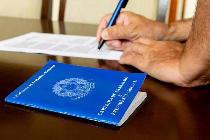 Mãos de homem assinando contrato ao lado de uma carteira de trabalho