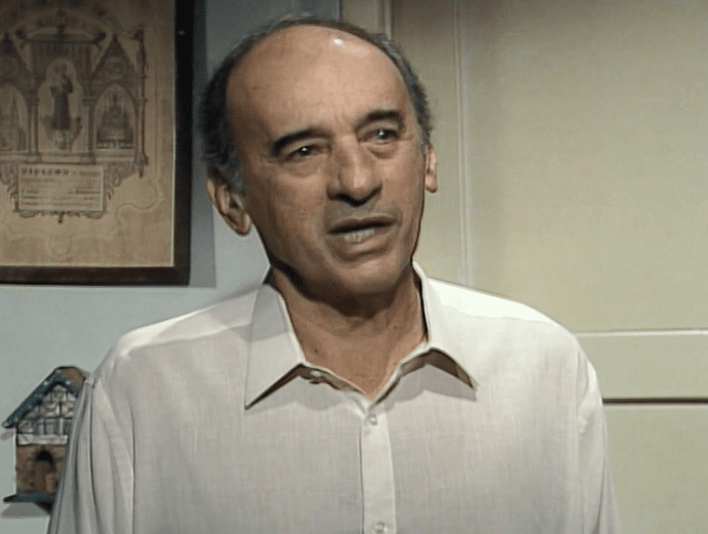 Personagem Salvador em Vale Tudo, homem mais velho, de pele clara, cabelos ralos e escuros nas laterais, aparece em ambiente interno usando uma camisa social clara. Ele está em discussão com a personagem Maria de Fátima. Ao fundo, vê-se uma parede com um quadro de moldura escura e uma pequena prateleira com um objeto decorativo em forma de casinha.